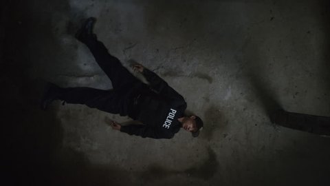Overhead shot of fallen police officer lying on a concrete floor in underground tunnel revealing special ops military SWAT team members approaching body with flashlights and guns drawn, then getting s