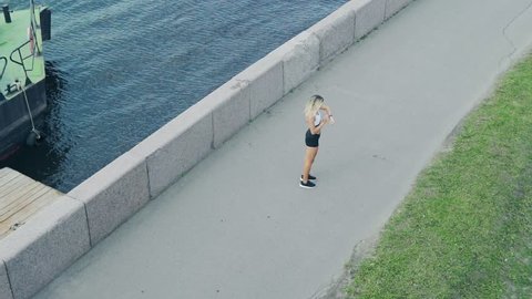 Above view of blonde female athlete running in slow motion along embankment next to river, stopping to check settings on her fitness tracker and proceeding marathon training