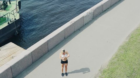High angle of female athlete running along embankment next to river, stopping to check distance and speed on her smartwatch and proceeding marathon training
