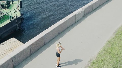 High angle of fit woman jogging along embankment next to river, stopping to check fitness tracker on her wrist and proceeding race