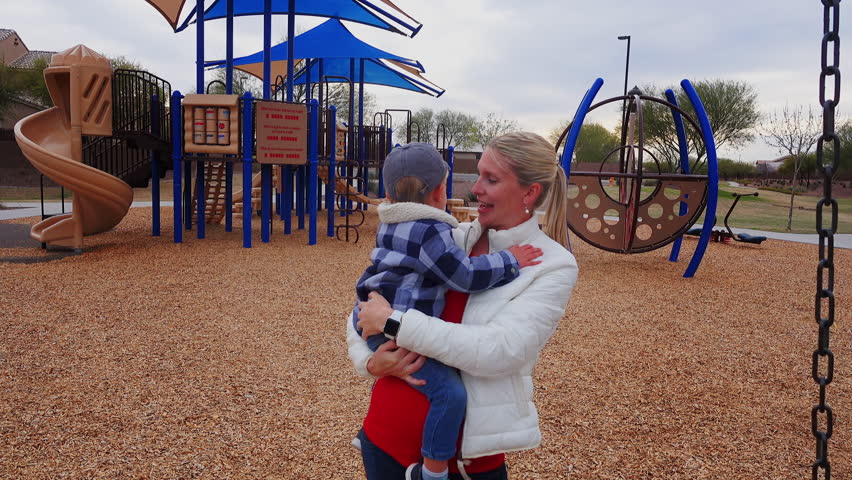 A young mother holds her son and waves to the camera in a typical Arizona neighborhood playground on a chilly winter day. Phoenix suburbs.  	