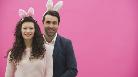 Young couple standing on a pink background. During this, the gesture class shows a smile, looking at the camera. Happy family is preparing for Easter, with the ears of a pink rabbit on its head.