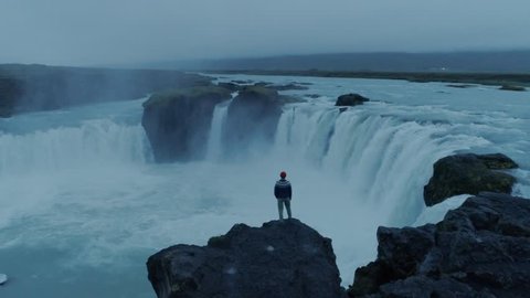 Aertial footage of drone fly around confident, brave and independent contemporary man, hipster intellectual nomad traveller stand on cliff or edge of waterfall in dusk, look ahead for new adventures