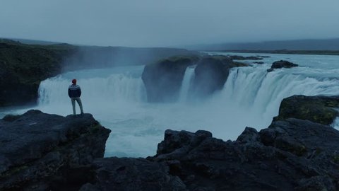Drone camera flies around hipster young man in blue knit wool sweater and red beanie, stand on edge of cliff or rock overlooking epic mountain icelandic waterfall on moody summer night light