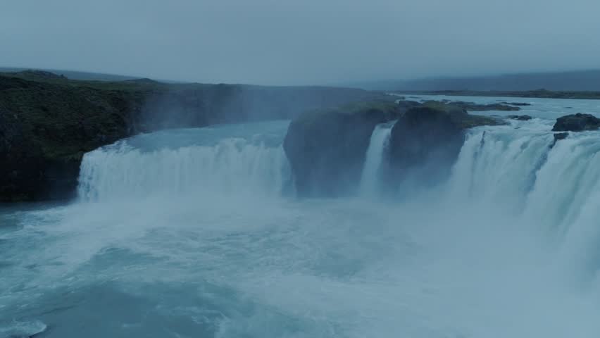 Cinematic drone shot of bikepacker or traveller sit on edge of cliff or rock, observe waterfall on cloudy and moody day. Lonely and sad, thoughtful pensative deep in his thoughts connects with nature