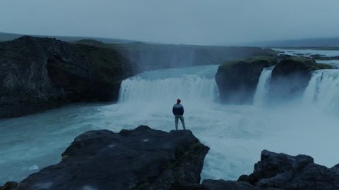 Epic adventure seeker drone shot of urban nomad traveller,hipster young millennial man stand on edge of cliff near waterfall in iceland, confident and brave, search for new exciting horizons and ideas