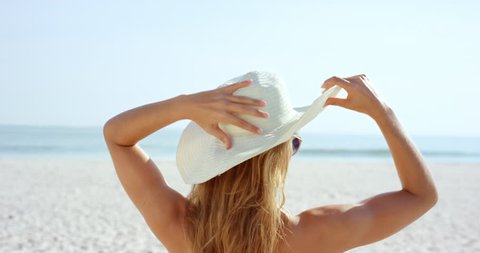 beautiful young woman putting on sun hat at beach wearing white swimsuit