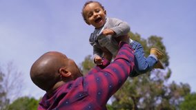 African-american young bald father in striped shirt and jeans throwing up his little mixed-race boy into air and catching him into hands, child laughing. Family, holiday concept
