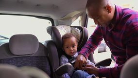 Tired little mixed-race boy sitting in baby seat, his African-american young bald father in striped shirt and jeans fastening seat belts. Family, safety concept