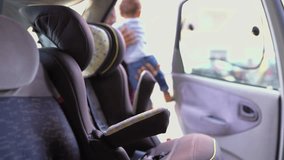 Afro-american young bald father in striped shirt and jeans putting his little mixed-race boy into baby seat, fastening seat belts. Side view. Family, safety concept