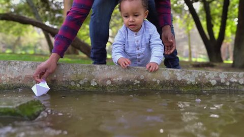 Little mixed-race boy in park playing with water in fountain with African-american father in striped shirt and jeans, father sailing off paper ship sailing off, son catching it. Family, game concept