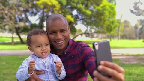 Afro-american young bald father in striped shirt and jeans sitting on haunches in park, holding his little mixed-race son on knees, making selfie. Front view. Family, holiday concept