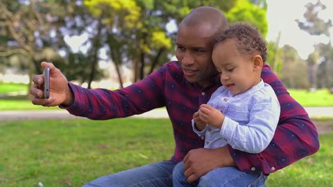 African-american young bald father in striped shirt and jeans sitting on haunches in park, holding his little mixed-race son on knees, making selfie. Side view. Family, holiday concept