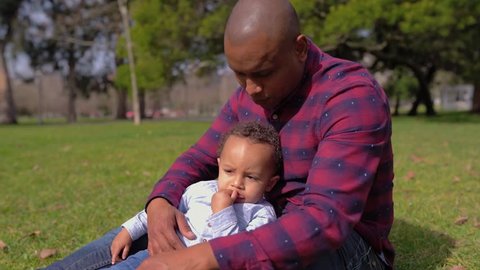 Afro-american young bald father in striped shirt and jeans sitting on grass in park, holding his little mixed-race son on knees, hugging. Family, holiday concept