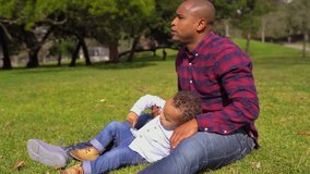 Afro-american young bald father in striped shirt and jeans sitting on grass, raising up his little mixed-race boy from grass, making him seated on knees, boy looking tired. Family, weekend concept