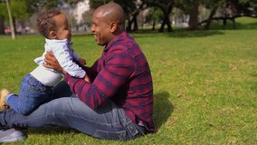 Afro-american young bald father in striped shirt and jeans sitting on grass, playing with his little mixed-race boy, holding him in his arms. Family, weekend concept