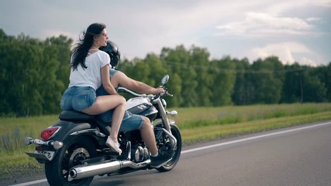 young couple in denim summer dressing rides black metal modern motorbike along gray road under cloudy sky