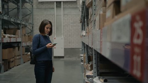 Young Caucasian woman with smartphone choosing goods in shop warehouse.
