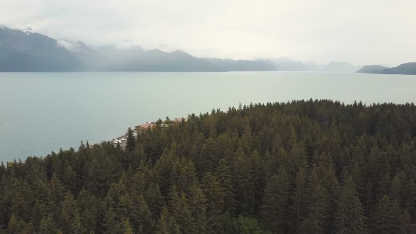 Aerial over Alaskan forest towards seaside highway with snowy mountains in distance, Seward, USA