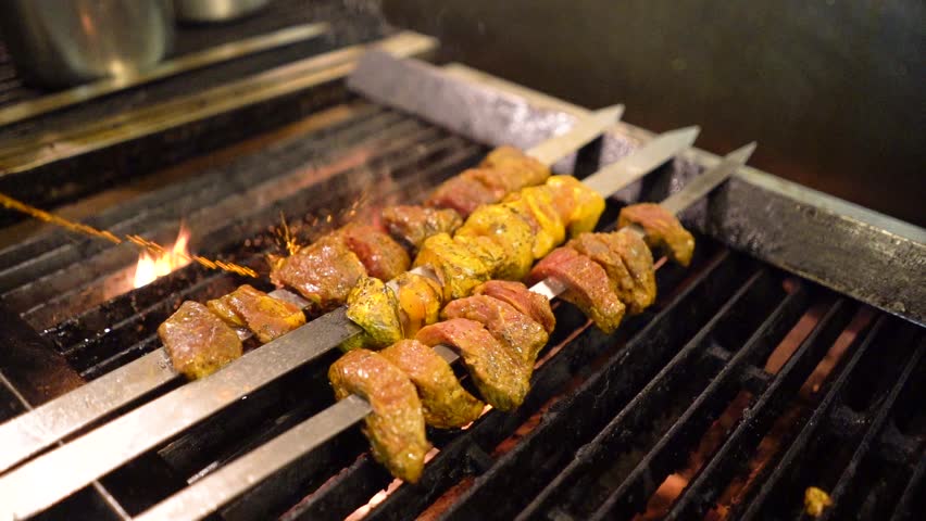 Slow motion of skewers of lamb, beef and chicken kabobs on a grill in a kitchen with flames flaring up.