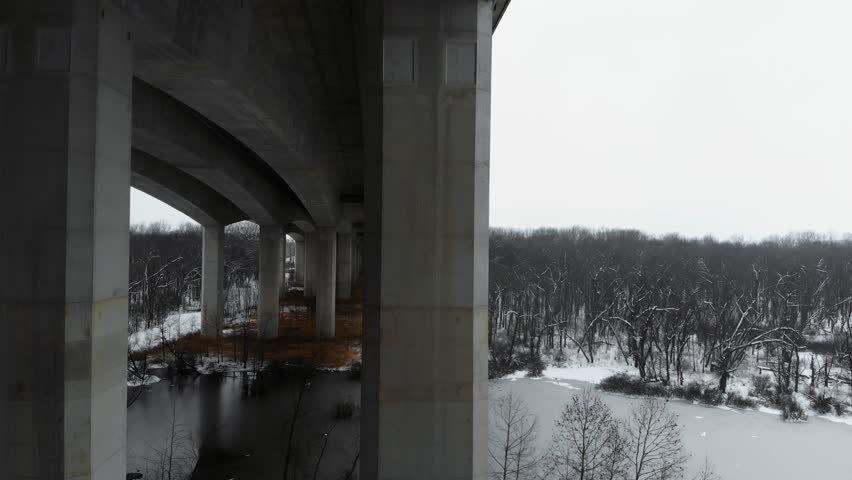 A drone flies under Pace Bridge in St. Louis, Missouri over a snow-lined lake.