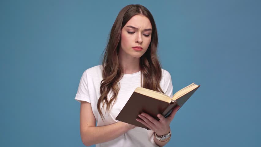 beautifu confused girl reading book, looking away and raising eyebrows isolated on blue