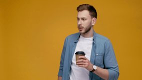 handsome man drinking coffee to go, looking at camera, smiling and showing paper cup at camera isolated on yellow