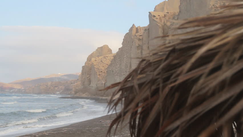 Detail of a straw beach umbrella with the black beach and white cliff formations in the background. Waves hit the beach. Santorini, Greece.