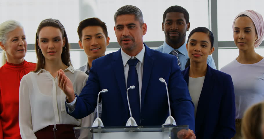 Front view of a Caucasian businessman speaking to the public while standing on the podium with his colleagues in the business seminar.