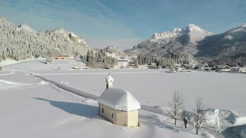 Aerial Drone shot showing a snowy landscape in in the sunshine of the Tyrol, Austria and a snow covered tiny chapel in Lechaschau.