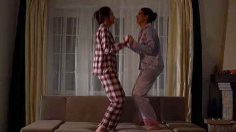 Two cheerful happy sisters twins in pajams jumping on the couch in a cozy living room and having fun like in childhood. Relationship sisters. Slow motion.