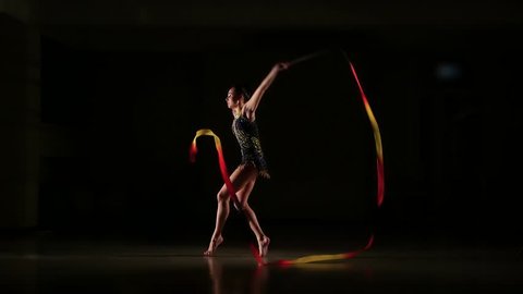 Slow motion, slim cute attractive girl athlete in bright colorful swimsuit performs elements of rhythmic gymnastics with ribbon in dark gym with beautiful staged light