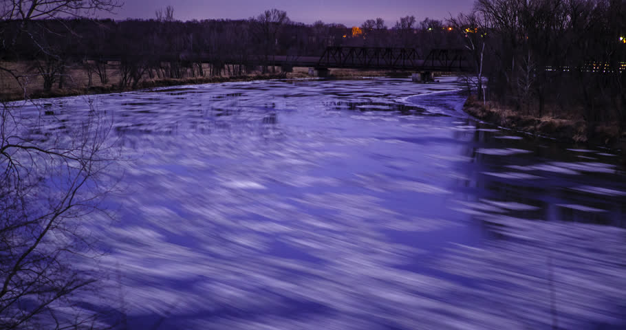 Rapidly flowing river freezing over to a sudden stop under moon and starlight with a freight train passing over bridge in background. 
