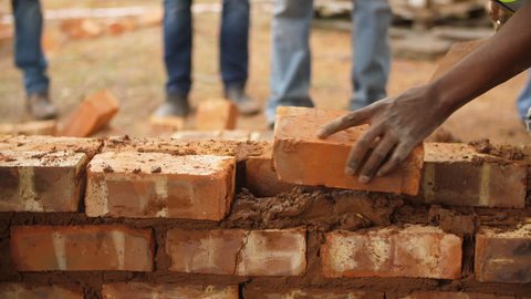Black male volunteer worker lays brick into wall with cement using trowel