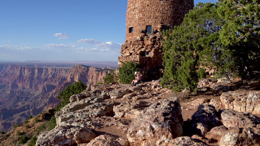 tall watch tower located on the top viewpoint of desert mountain in Grand Canyon National Park arizona usa. two young female hikers resting on wooden bench chair under sunshine blue sky near the tree