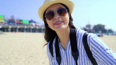 cheerful young asian woman backpacker relaxing on the seaside beach bay on Santa Monica Pier california. girl traveler with hat and sunglasses waving hands talking on video phone call showing view.