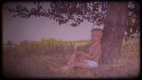 Family video archive. Retro camera 8 mm. Vintage. A little boy sits and dreams under a big tree. Childhood dreams. Place of solitude. Journey back to childhood. Fabulous tree. Memories.  A warm wind