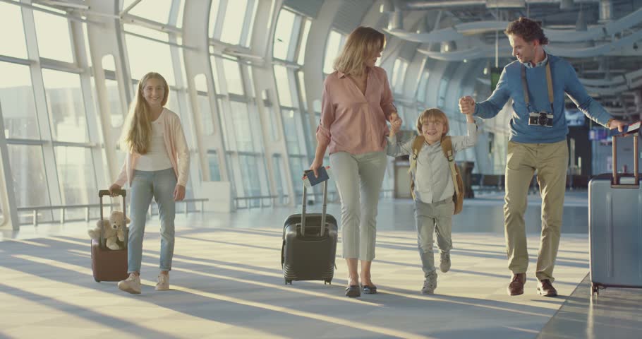 Caucasian happy mother, father, little daughter and son walking the airport hall and carrying their suitcases on the wheels before the departure to the vacation and laughing cheerfully 
