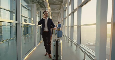 Caucasian handsome man in business style stepping tothe boarding through the airport corridor, carrying suitcase with wheels and talking on the phone, businesswoman following him.