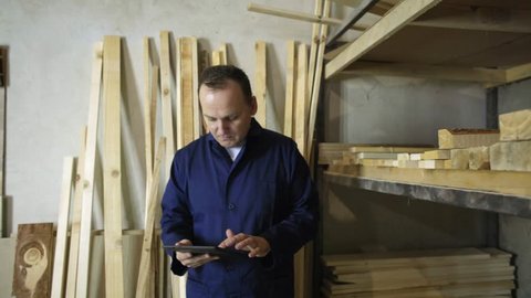 A man with a tablet is walking in a furniture factory and checking up the wooden stack 
