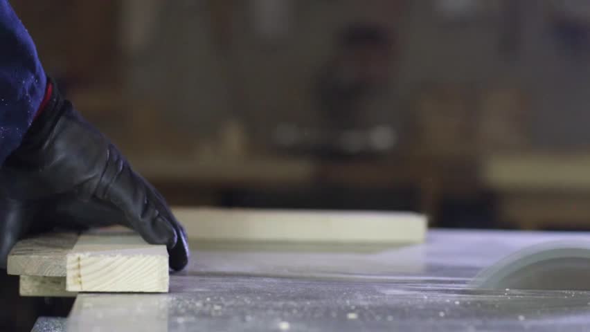 Process of furniture production. A young man in a dark coat cutting the wooden pieces for the sofa on a cutting machine. He is wearing a safety glasses and gloves.
