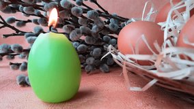 Looped video: burning green candle made in shape of easter egg next to the willow branches with buds, brown chicken eggs in basket with straw