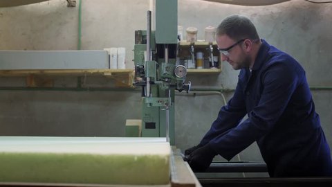 Process of furniture production. A young man in a dark coat cut the foam for the sofa on a cutting machine. Behind him there are a lot of finished sofas. He is wearing a safety glasses and gloves.
