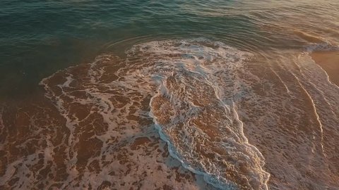 Drone aerial flying above a beautiful coastal shoreline with orange colored sandy beach, crystal clear blue crashing waves and a golden horizon at sunrise on a bright sunny day in Hawaii