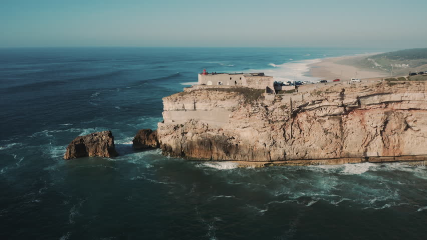 Aerial; drone flight over the Fort in Praia do Norte located on rocky slope; one of the most popular seaside resorts; biggest waves ever surfed; touristic attraction, long wide sandy beach viewpoint Royalty-Free Stock Footage #1026036767