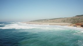 Aerial view; drone shot of lond wide sandy beach with curling crushing waves ; sunny weather in Nazare, Portugal; wind turbine located on background; windy and wavy beach with surf camp; nature power