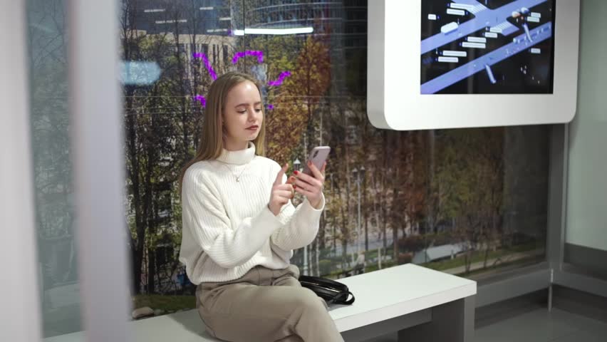 Young woman waiting and talking by phone at glass bus stop