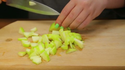 A woman cuts a green apple with a knife on a wooden board into small cubes.