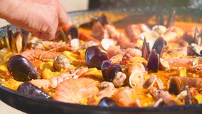 Paella traditional Spanish food, seafood paella in the fry pan with mussels, king prawns, langoustine and squids. Person cooking paella outside. Slow motion 4K UHD video