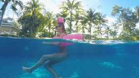 Playful young woman in resort swimming pool having fun with inflatable flamingo in tropical destination. Half in water shot of people paying while on vacations in beautiful hotels 